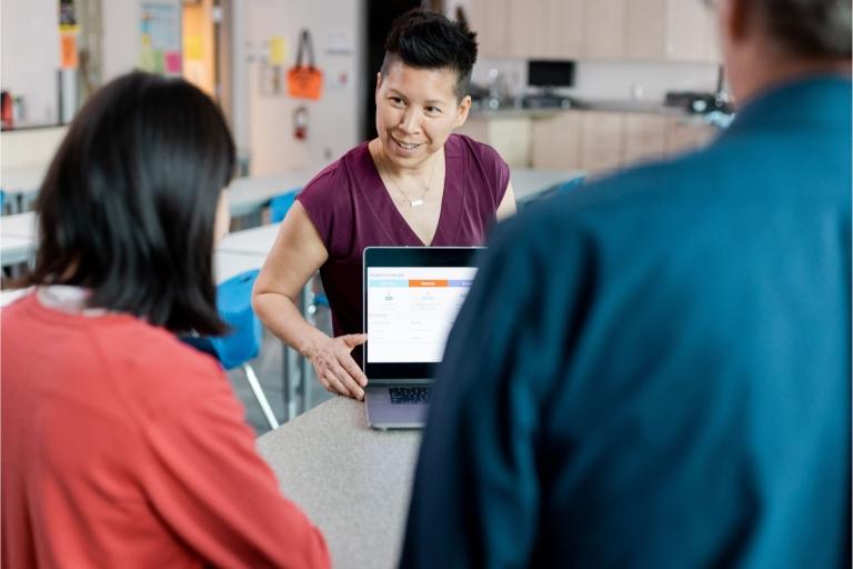 Teacher points to laptop in front of 2 students 